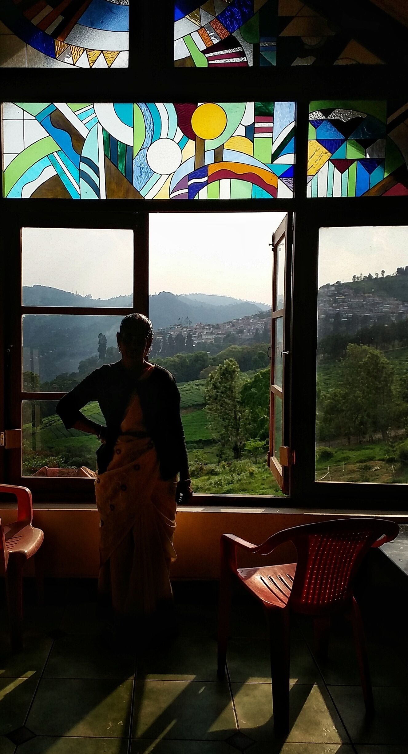 A person standing by a window with stained glass panes, overlooking a green landscape with hills.