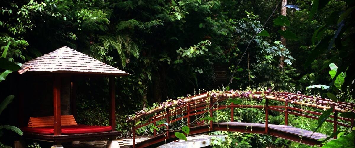 A wooden bridge with a red railing, a small gazebo, and lush greenery.