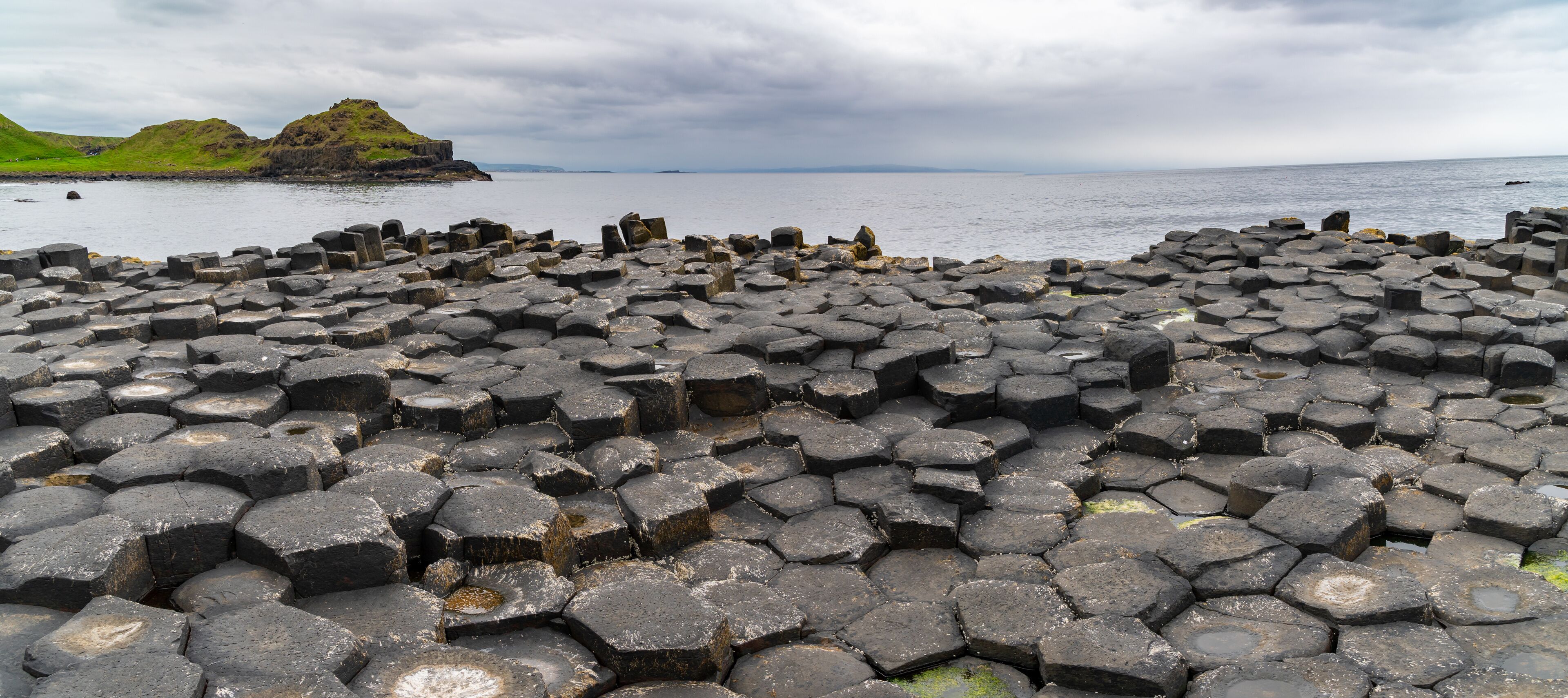 A rocky coastal area with hexagonal basalt columns and a distant green hill.