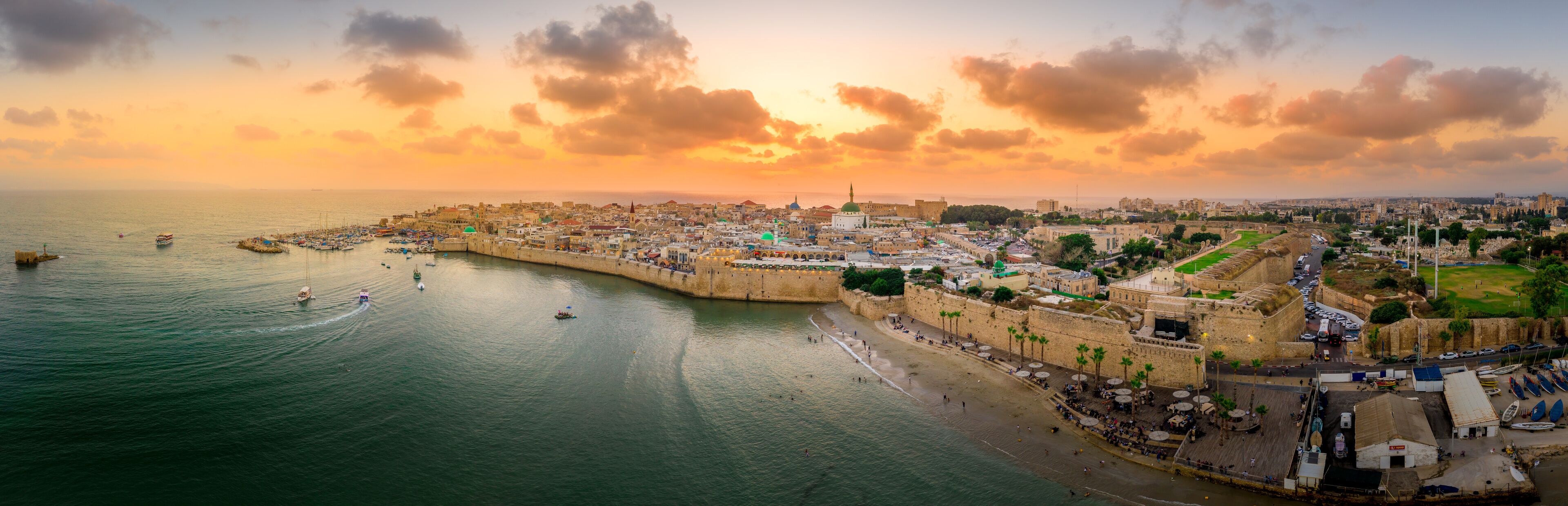 Un paisaje urbano costero con una fortificación histórica, un puerto deportivo y un cielo al atardecer.