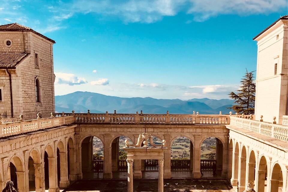Une cour intérieure ancienne, pavée de pierres, ornée d’arches et de statues, et offrant une vue sur des montagnes lointaines sous un ciel dégagé.