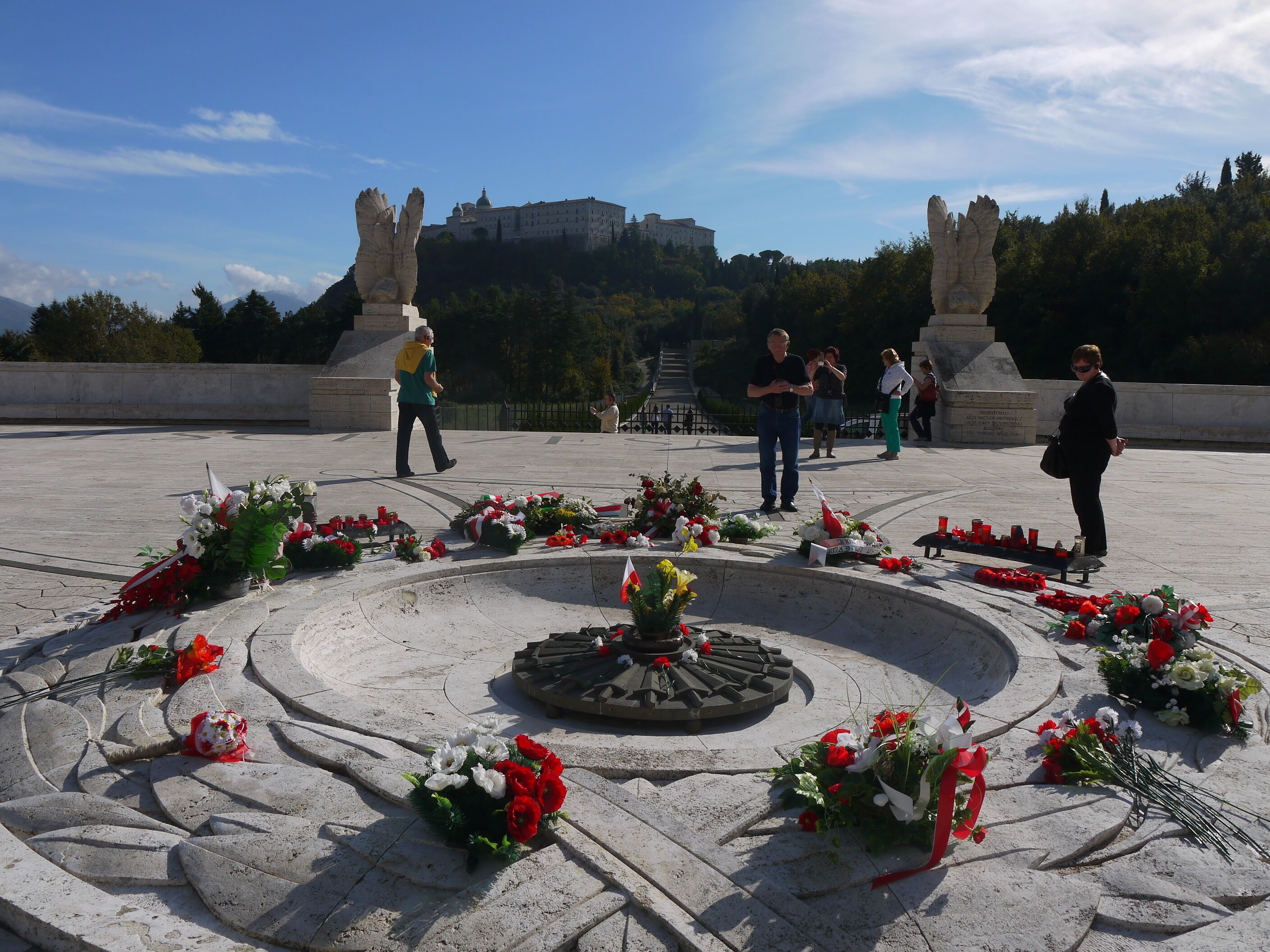 A memorial with flowers and a fountain, surrounded by a stone plaza with a castle in the background.