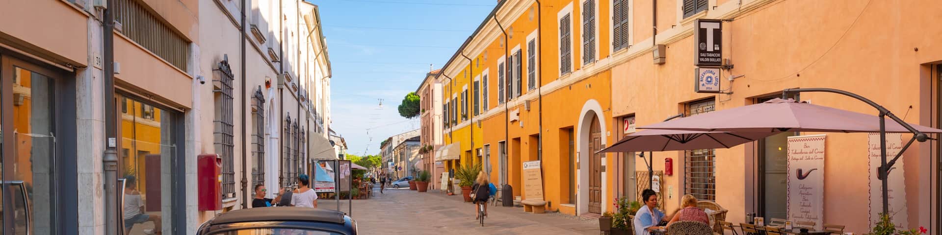 A narrow street with a vintage car parked on the side, shops, and outdoor seating at a cafe.