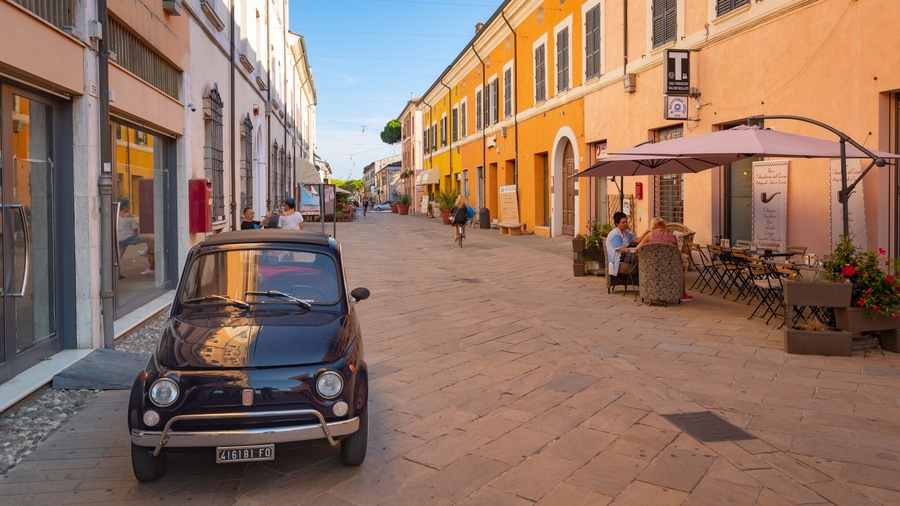 A narrow street with a vintage car parked on the side, shops, and outdoor seating at a cafe.