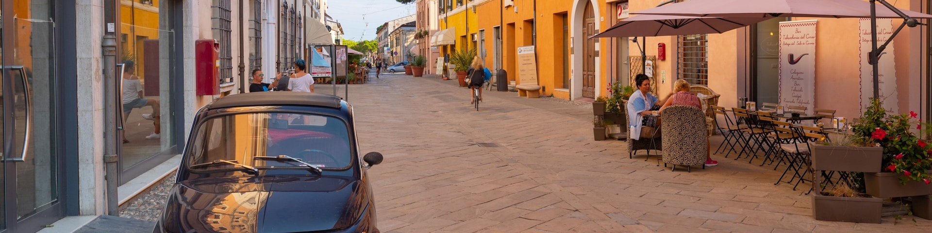 Una calle estrecha con un coche antiguo estacionado al lado, tiendas y mesas al aire libre en un café.