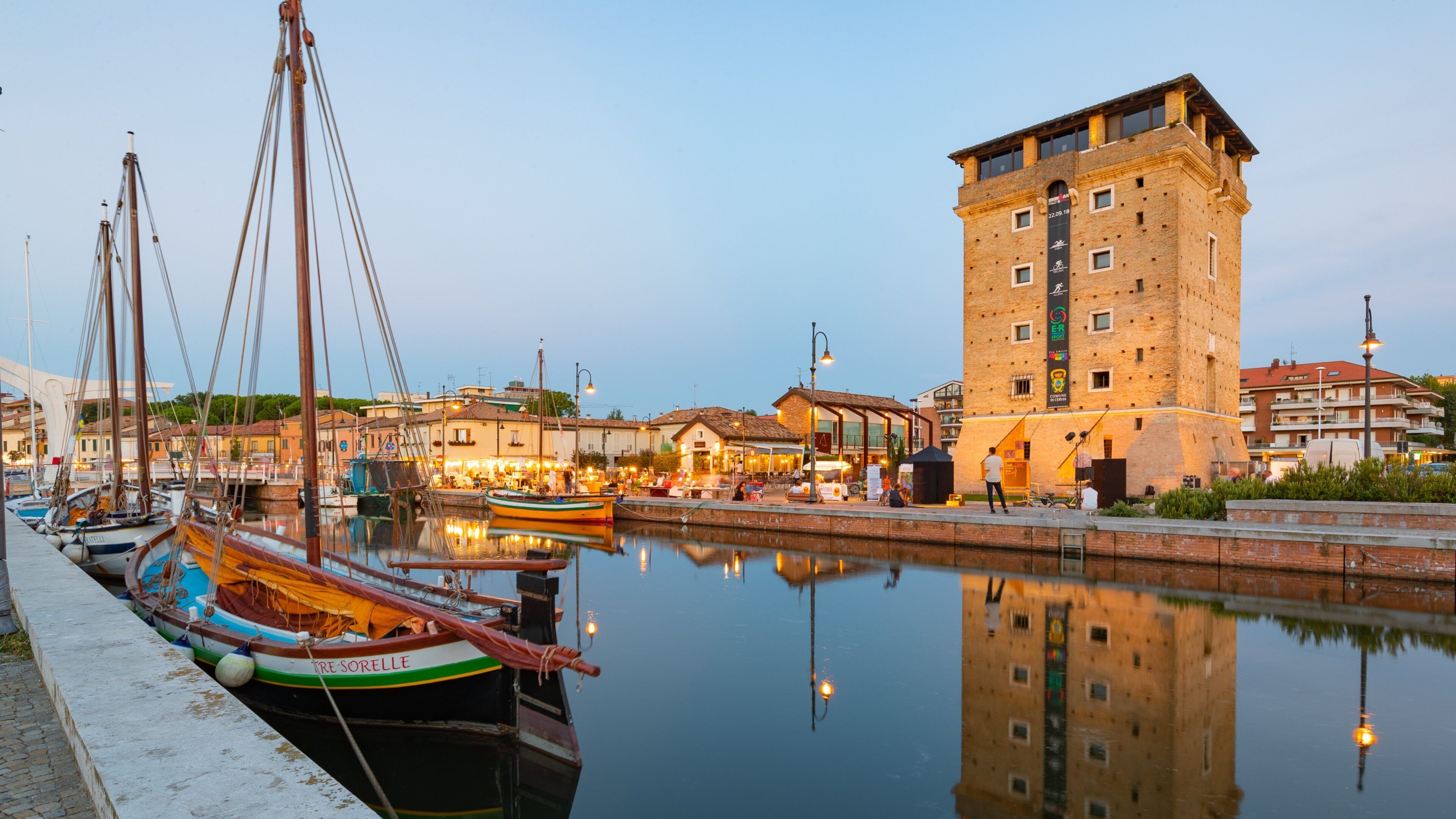 A harbor with boats docked, a tall brick building, and a calm water reflecting the surroundings.