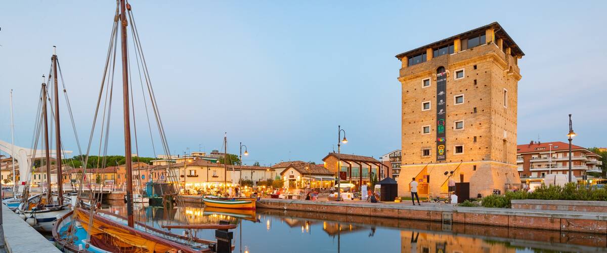 A harbor with boats docked, a tall brick building, and a calm water reflecting the surroundings.