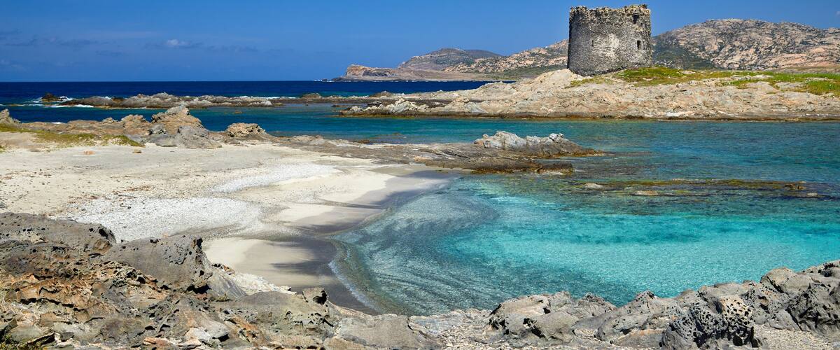 A coastal landscape with a rocky beach, clear turquoise waters, and a historic stone tower.