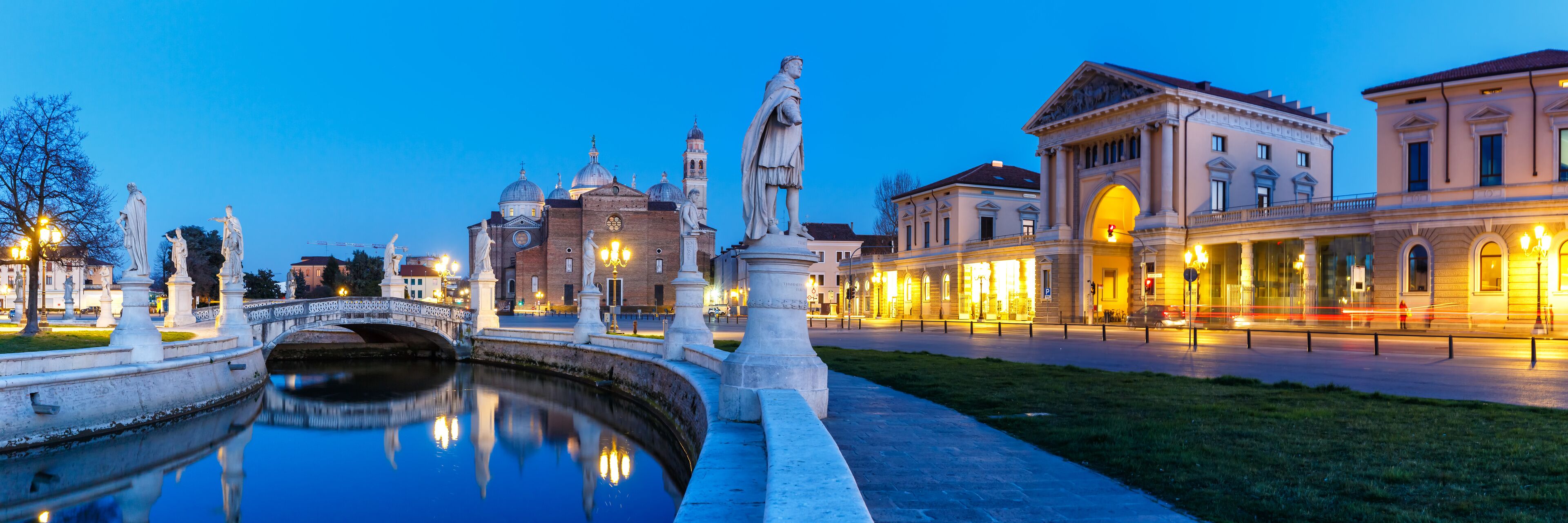A canal with statues, a bridge, and illuminated buildings at dusk.