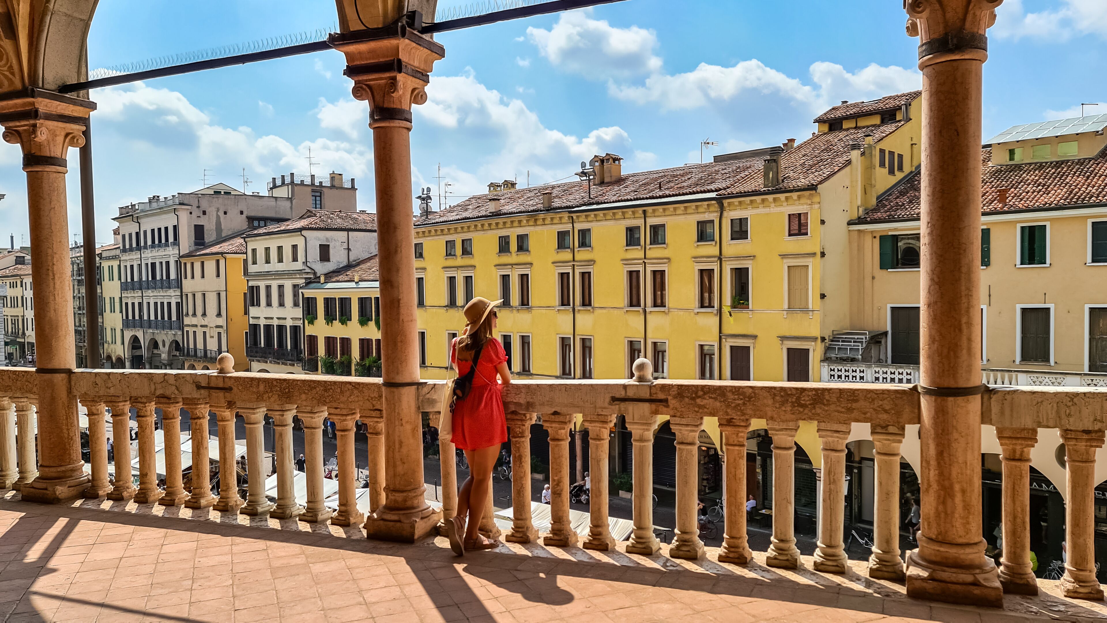 Tourist woman in red dress with scenic view on Piazza delle Erbe from loggia, external balcony of Palazzo della Ragione in Padua, Veneto, Italy, Europe. Light beams on vibrant colonnade, architecture