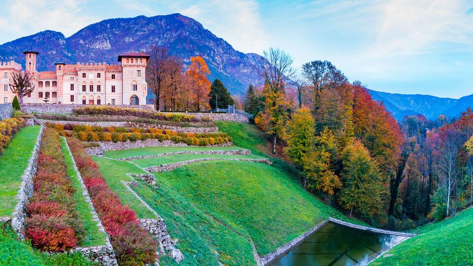 A castle with a tower, surrounded by autumn-colored trees and a mountainous backdrop.