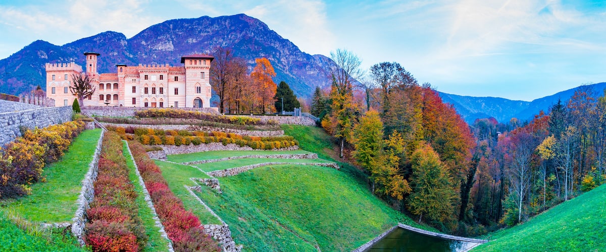 A castle with a tower, surrounded by autumn-colored trees and a mountainous backdrop.
