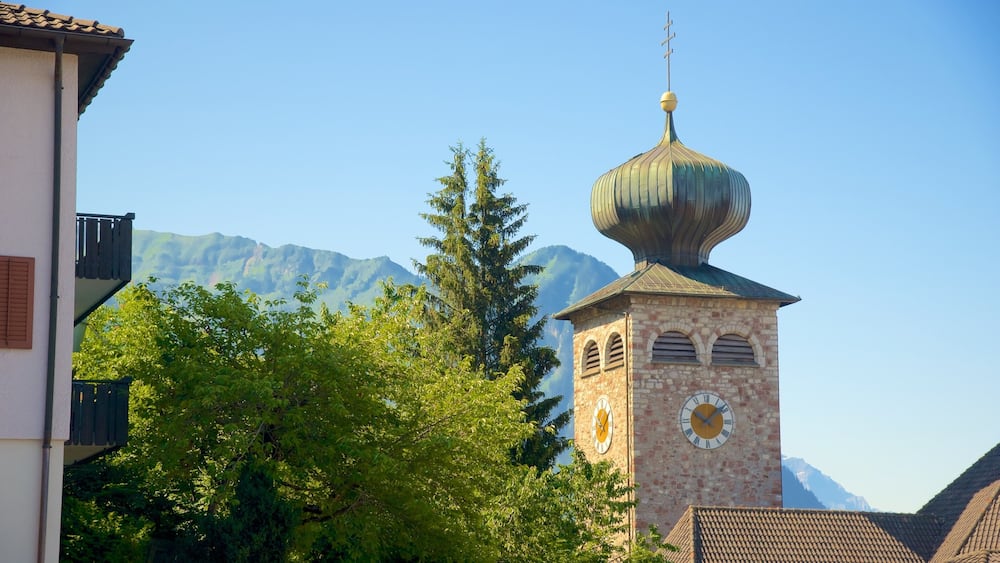 Un clocher d’église surmonté d’une horloge et d’un dôme, entouré d’arbres et avec des montagnes en arrière-plan.