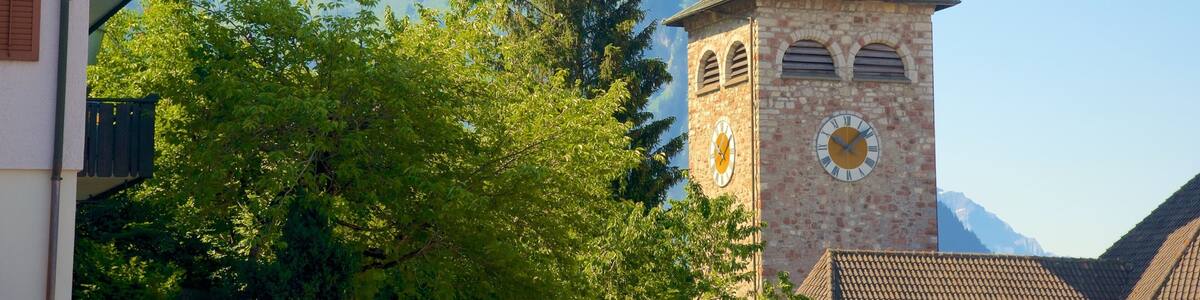 A church tower with a clock and a dome, surrounded by trees and mountains in the background.