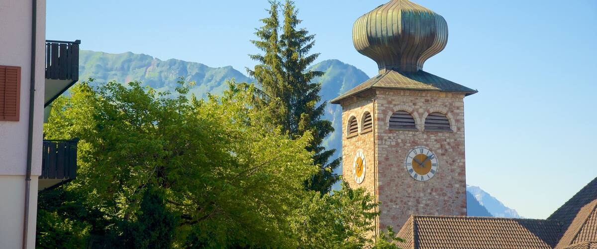 A church tower with a clock and a dome, surrounded by trees and mountains in the background.