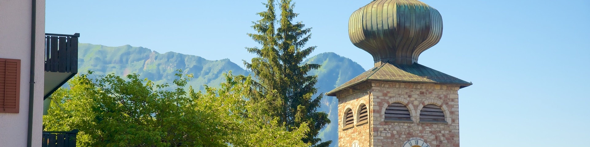 Una torre de iglesia con reloj y cúpula, rodeada de árboles y montañas al fondo.