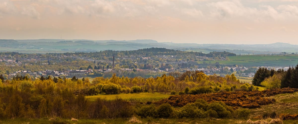 A panoramic view of a town nestled among rolling hills, with a dense forest in the foreground and a clear sky above.