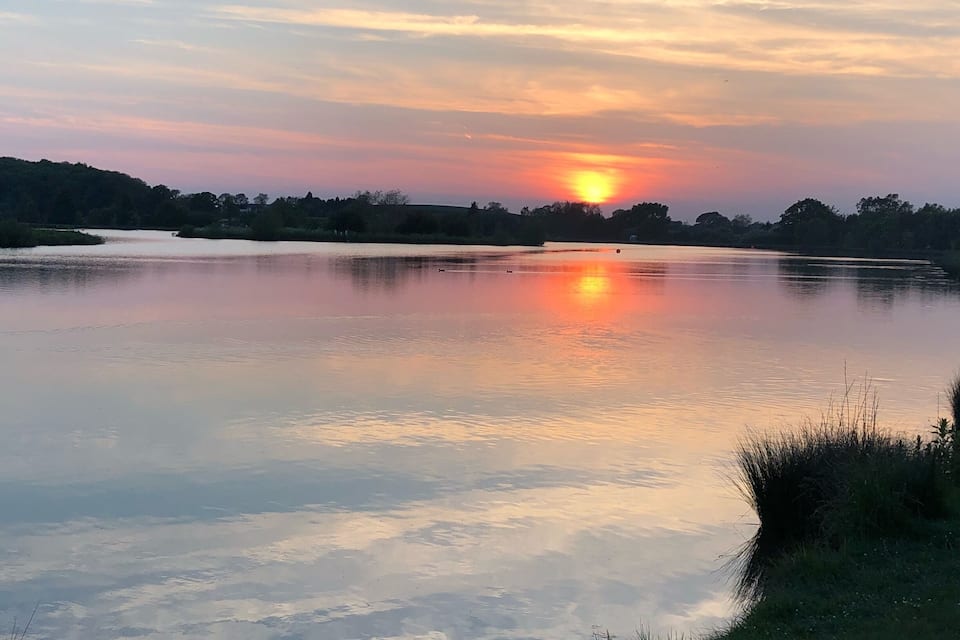 Un atardecer sobre un lago tranquilo, con reflejos del cielo, rodeado de árboles y orillas cubiertas de hierba.