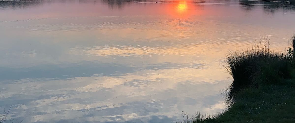 A sunset over a calm lake with reflections of the sky, surrounded by trees and grassy banks.