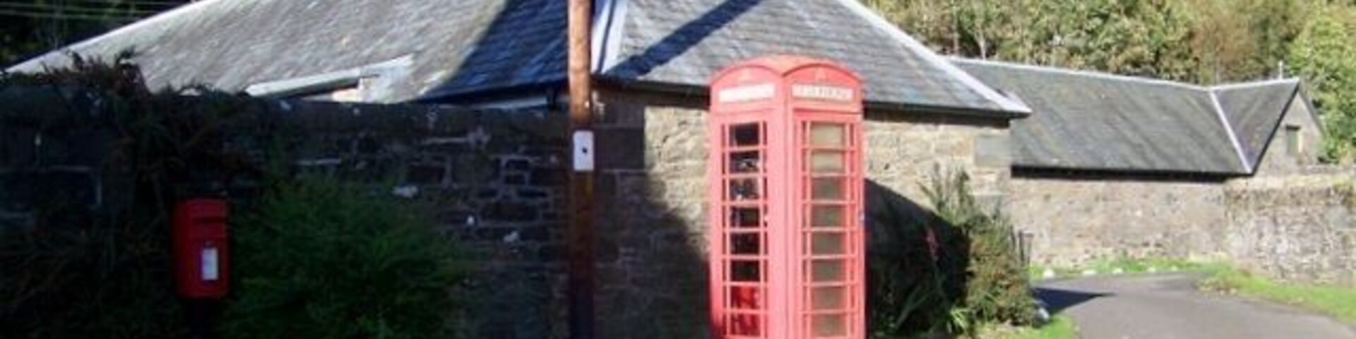 Una cabina telefónica roja, un edificio de piedra y una carretera en un entorno rural.