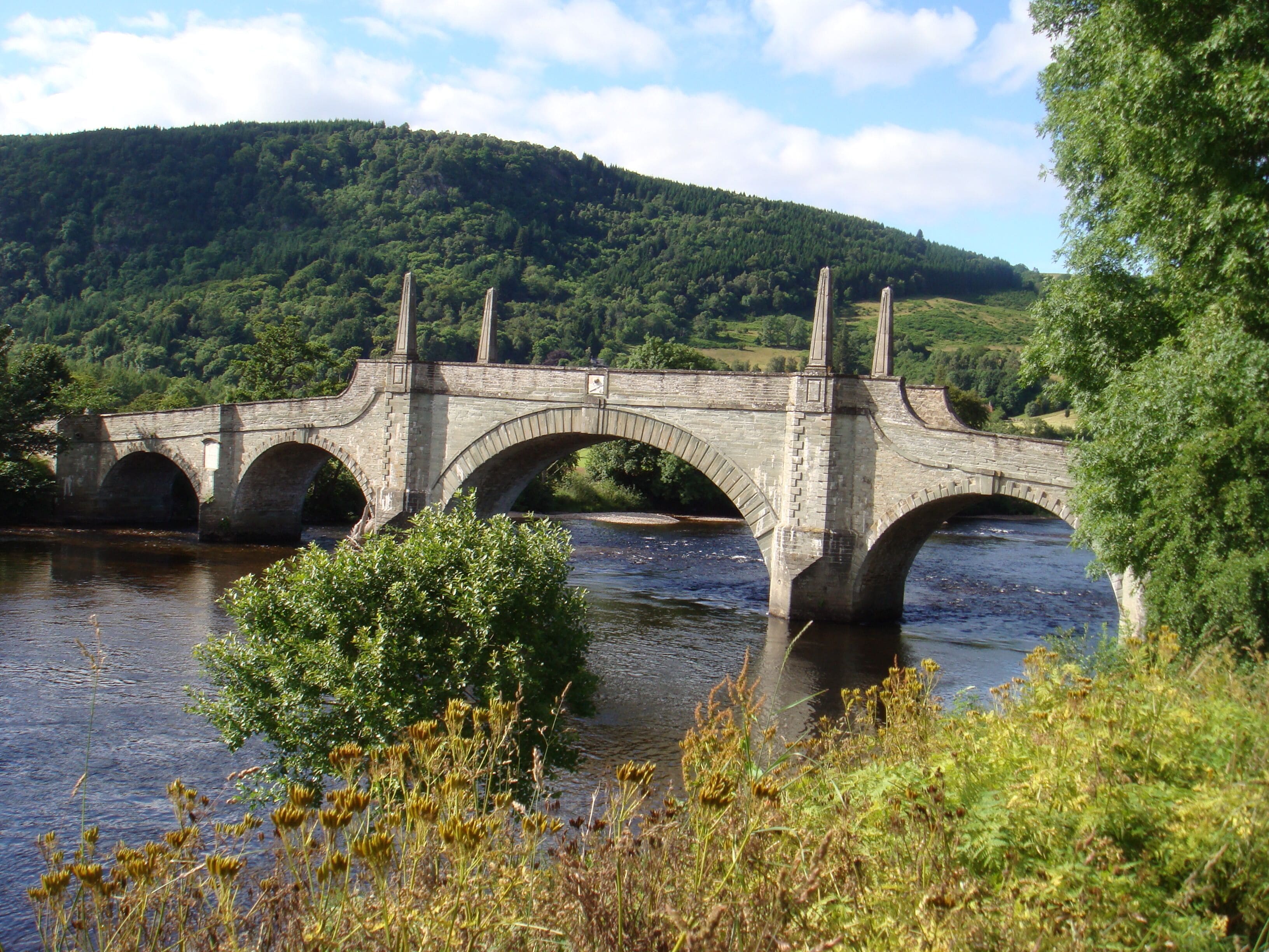 A stone bridge over a river with a forested hill in the background.