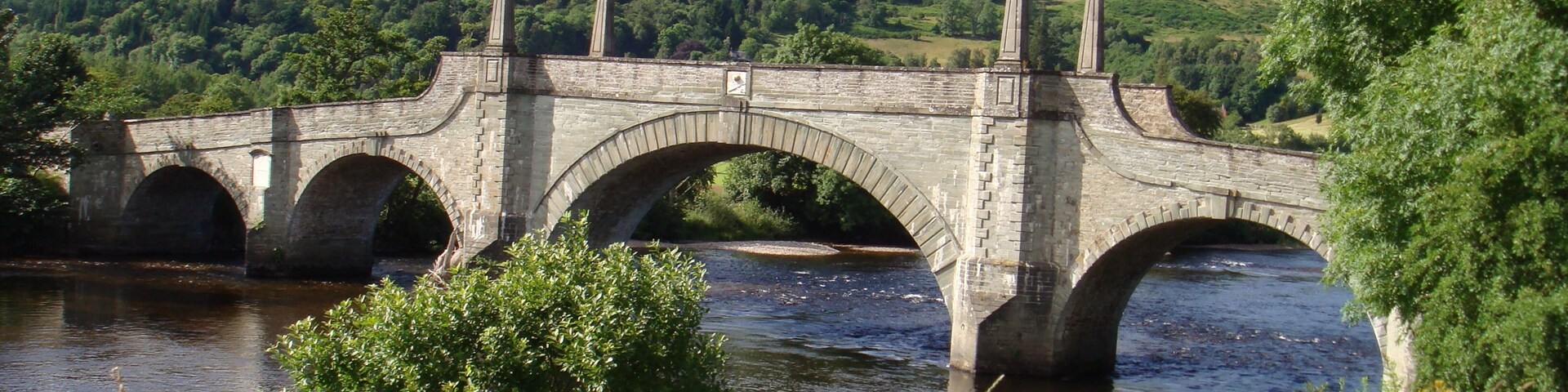A stone bridge over a river with a forested hill in the background.