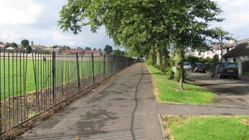 Ein gepflasterter Weg, gesäumt von Bäumen und einem Metallzaun, führt zu einer Wohngegend mit Häusern und geparkten Autos.