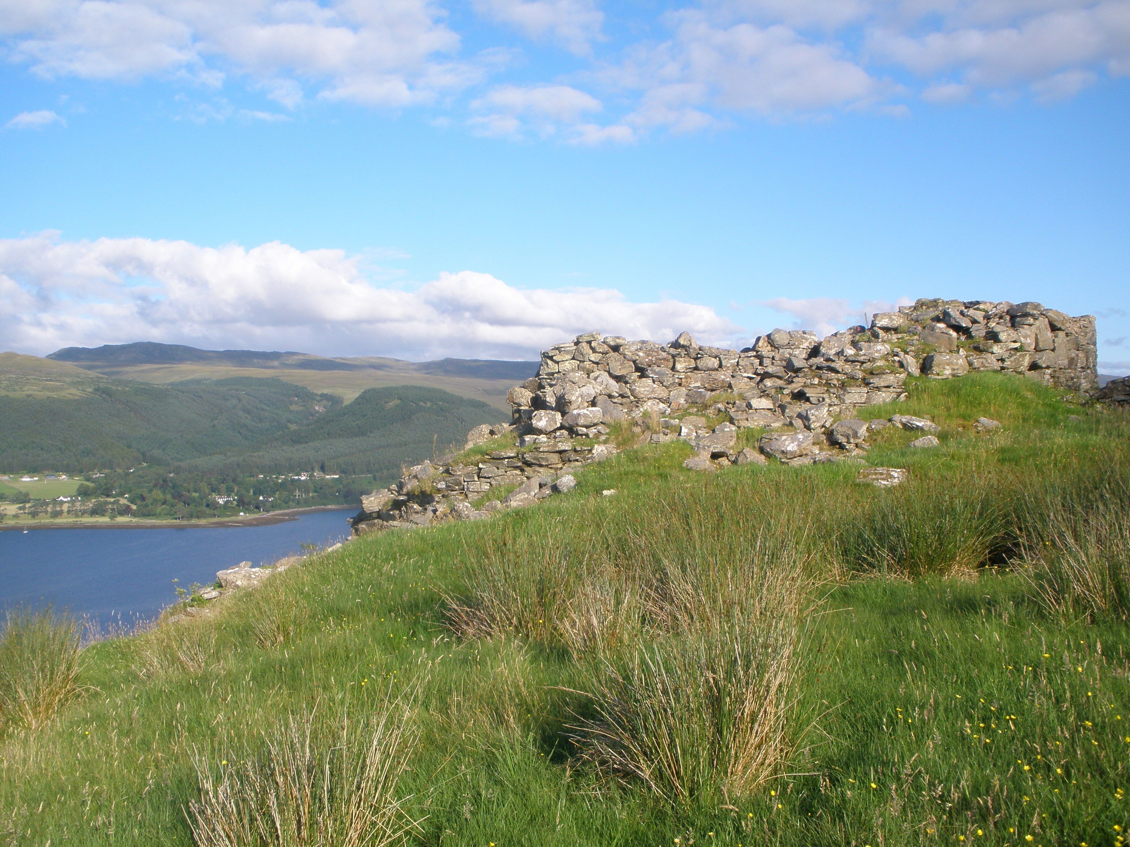 A stone ruin on a grassy hill overlooking a lake and mountains.