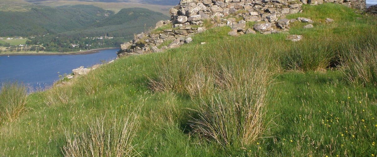 A stone ruin on a grassy hill overlooking a lake and mountains.
