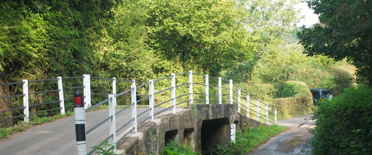 A narrow road passing under a stone bridge with metal railings and a red stop sign.