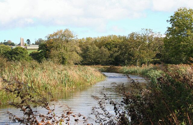 Burlescombe: Grand Western Canal. Behind the camera the canal has crossed a shallow valley on embankment running east-south-east; it turns to run north-north-east and in the distance is Burlescombe church. Had the original main line to Exeter been built, this is where it would have junctioned with the Tiverton branch. The Taunton-Exeter railway, which eclipsed the canal, runs nearby