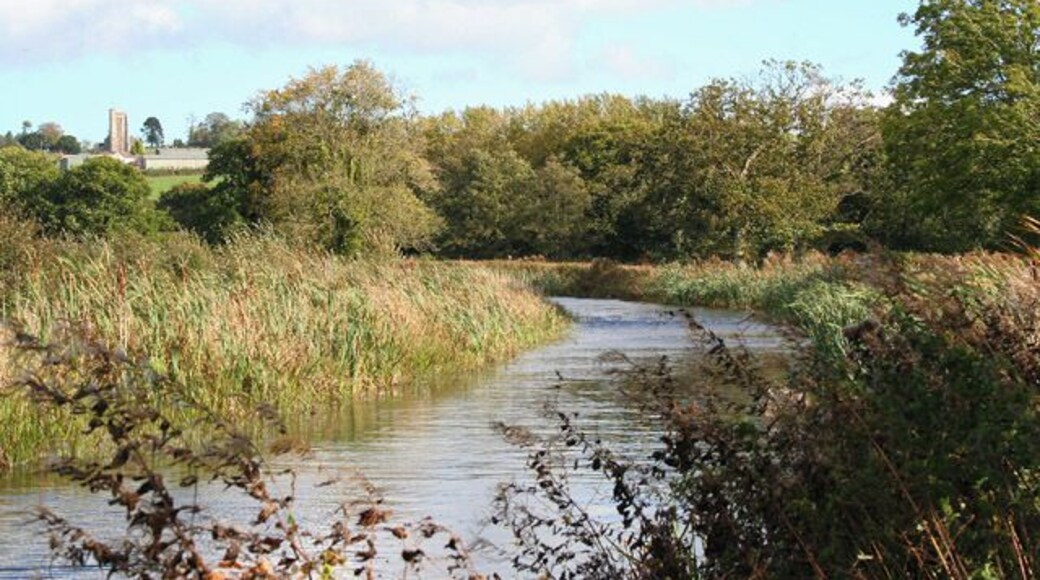 Burlescombe: Grand Western Canal. Behind the camera the canal has crossed a shallow valley on embankment running east-south-east; it turns to run north-north-east and in the distance is Burlescombe church. Had the original main line to Exeter been built, this is where it would have junctioned with the Tiverton branch. The Taunton-Exeter railway, which eclipsed the canal, runs nearby