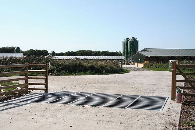 A farm entrance with a metal gate, a concrete pathway, and green silos in the background.