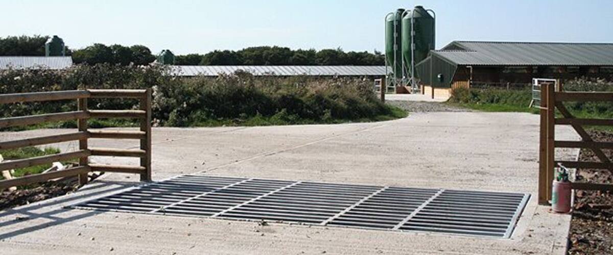 A farm entrance with a metal gate, a concrete pathway, and green silos in the background.