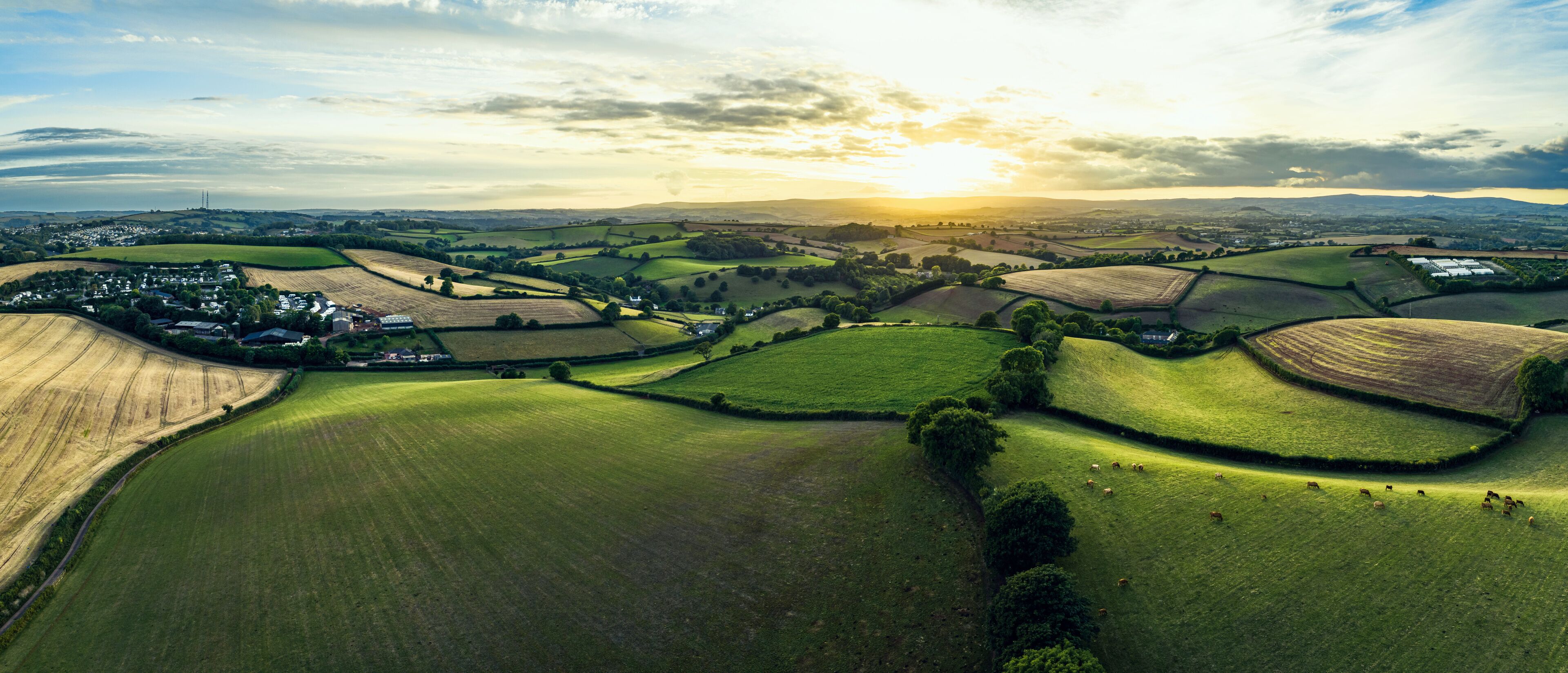 Rolling green fields, a village, and distant hills under a sunset sky.