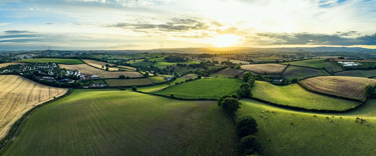 Rolling green fields, a village, and distant hills under a sunset sky.