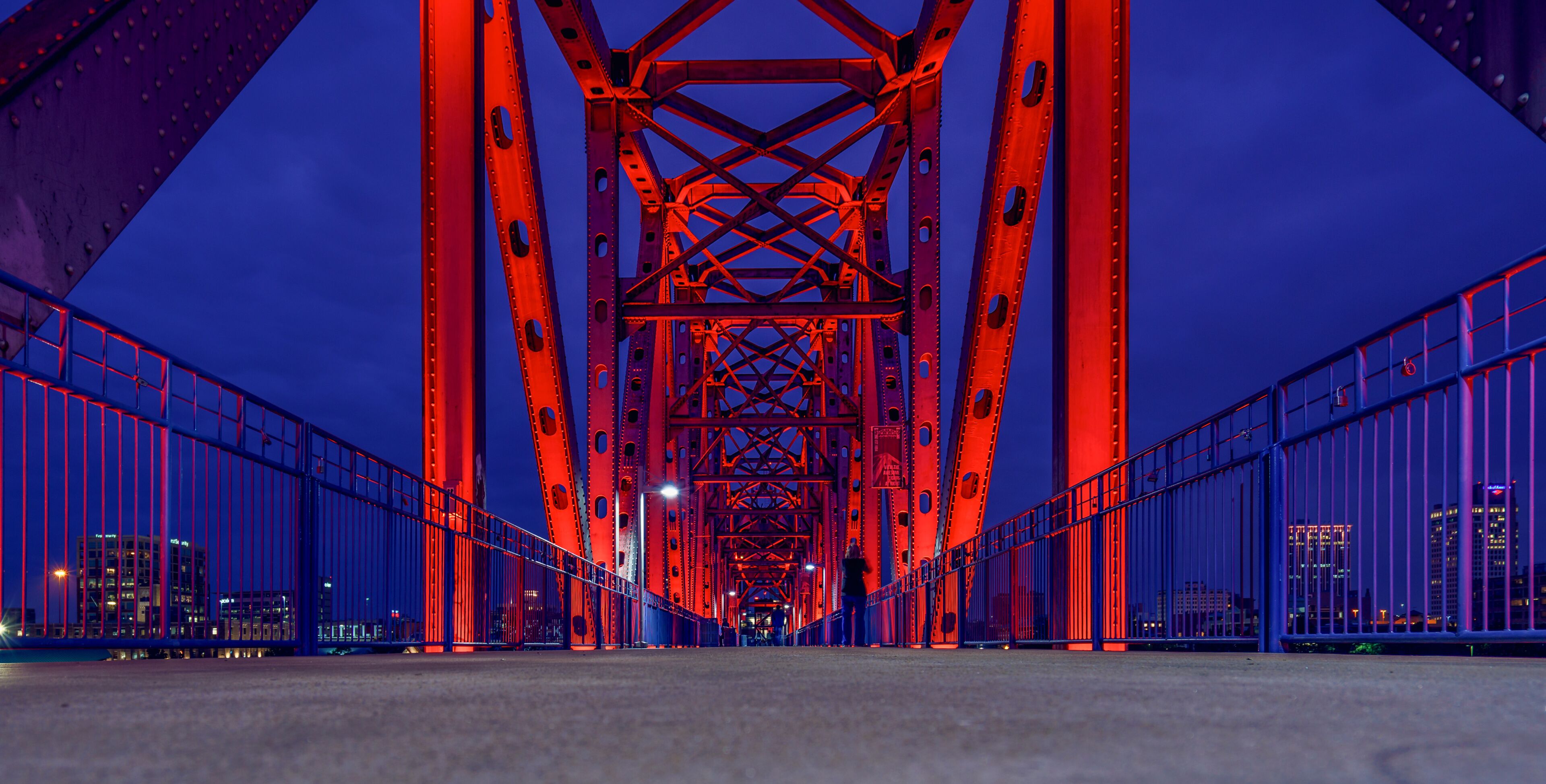 A nighttime view of a large red bridge with a complex truss structure, flanked by blue railings and city buildings in the background.