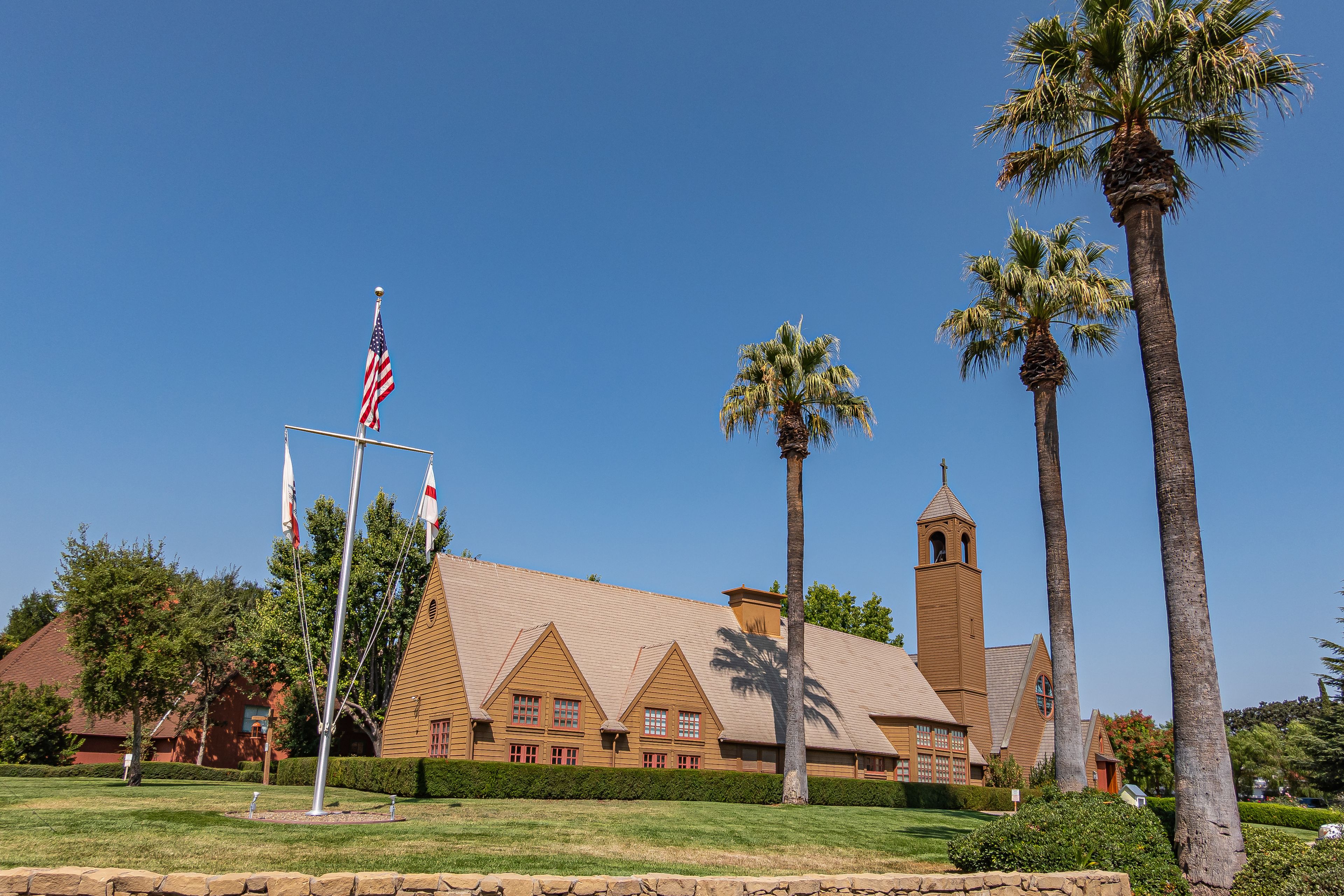 Los Olivos, California, USA - September 3, 2020: Wide view on brown wood St. Marks in the Valley Church with bell tower set in green park under blue sky, including side buildings. Flag post up front.