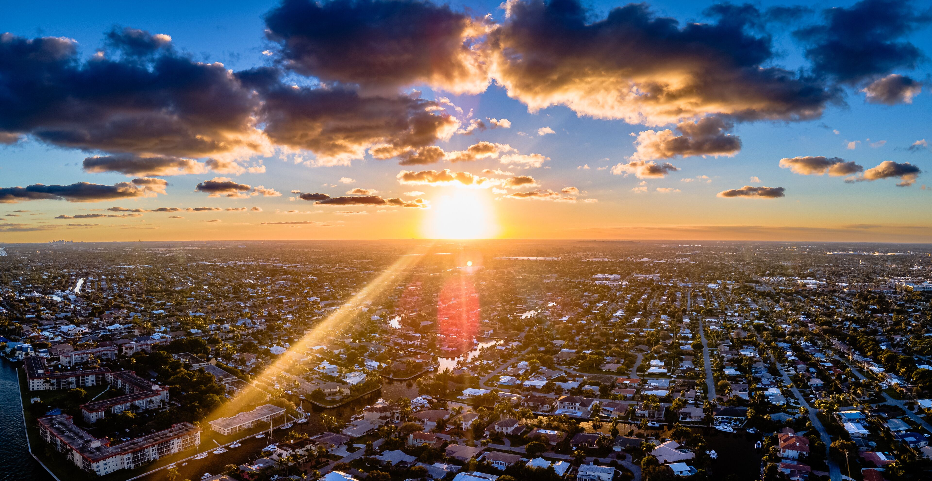 A cityscape at sunset with the sun low on the horizon, casting long shadows across the urban area.