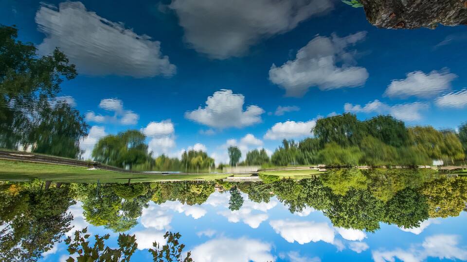 A clear blue sky with clouds, a calm body of water reflecting the sky, and dense green trees.