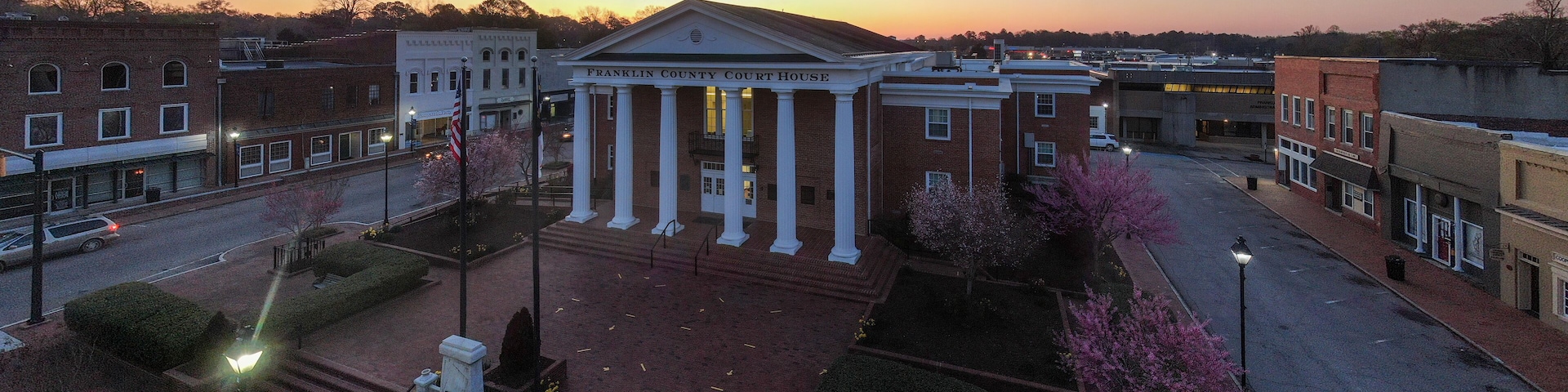 A courthouse with columns and a flag, surrounded by a landscaped area with trees and a street.