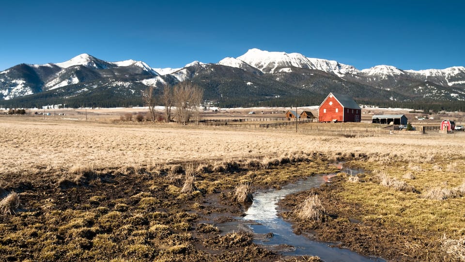 A rural landscape with a red barn, a muddy stream, and snow-capped mountains in the background.