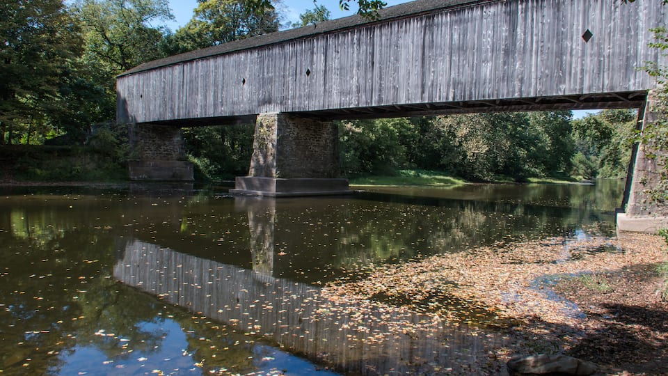 A wooden covered bridge over a calm river with reflections, surrounded by trees and a grassy bank.