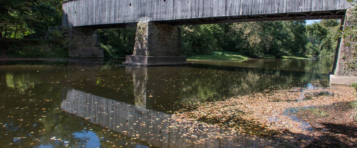 A wooden covered bridge over a calm river with reflections, surrounded by trees and a grassy bank.