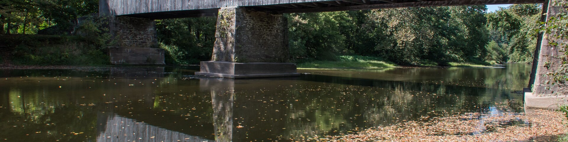 A wooden covered bridge over a calm river with reflections, surrounded by trees and a grassy bank.
