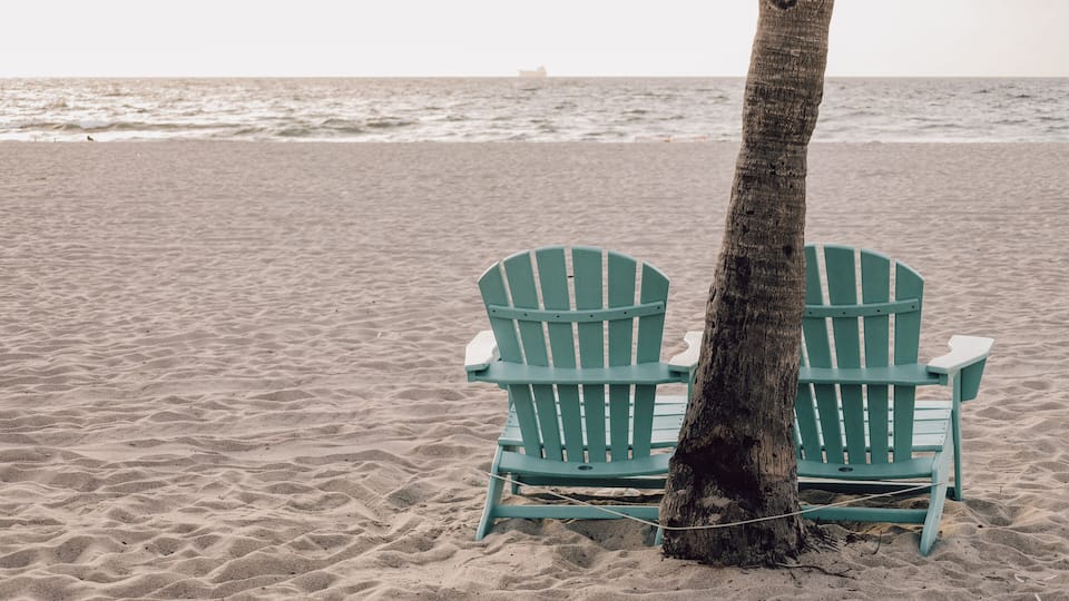 Two green Adirondack chairs tied to a tree on a sandy beach with the ocean and a ship in the distance.