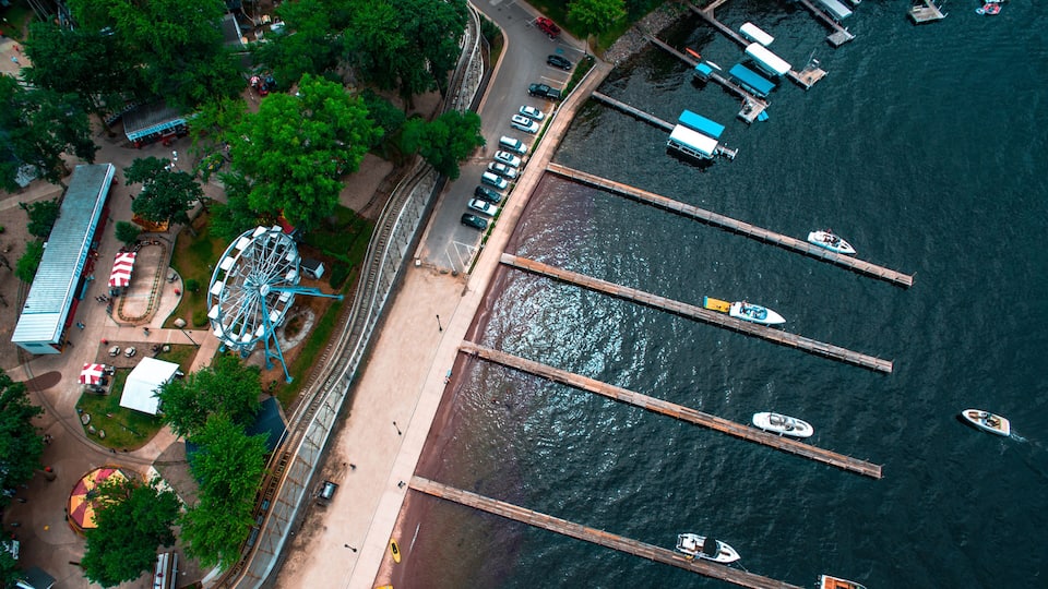 An aerial view of a waterfront area with a Ferris wheel, a parking lot, and several boats docked at piers.
