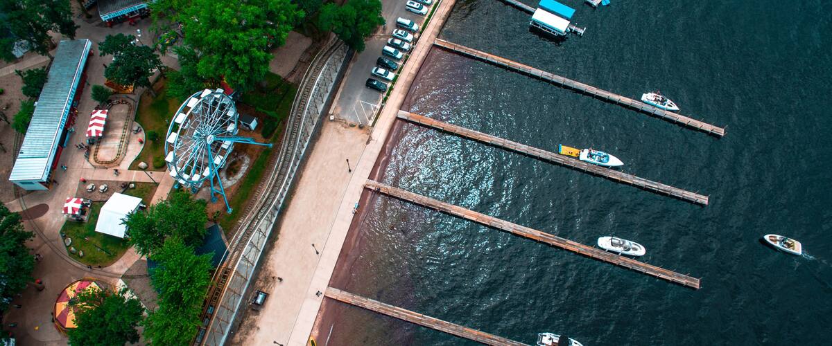 An aerial view of a waterfront area with a Ferris wheel, a parking lot, and several boats docked at piers.
