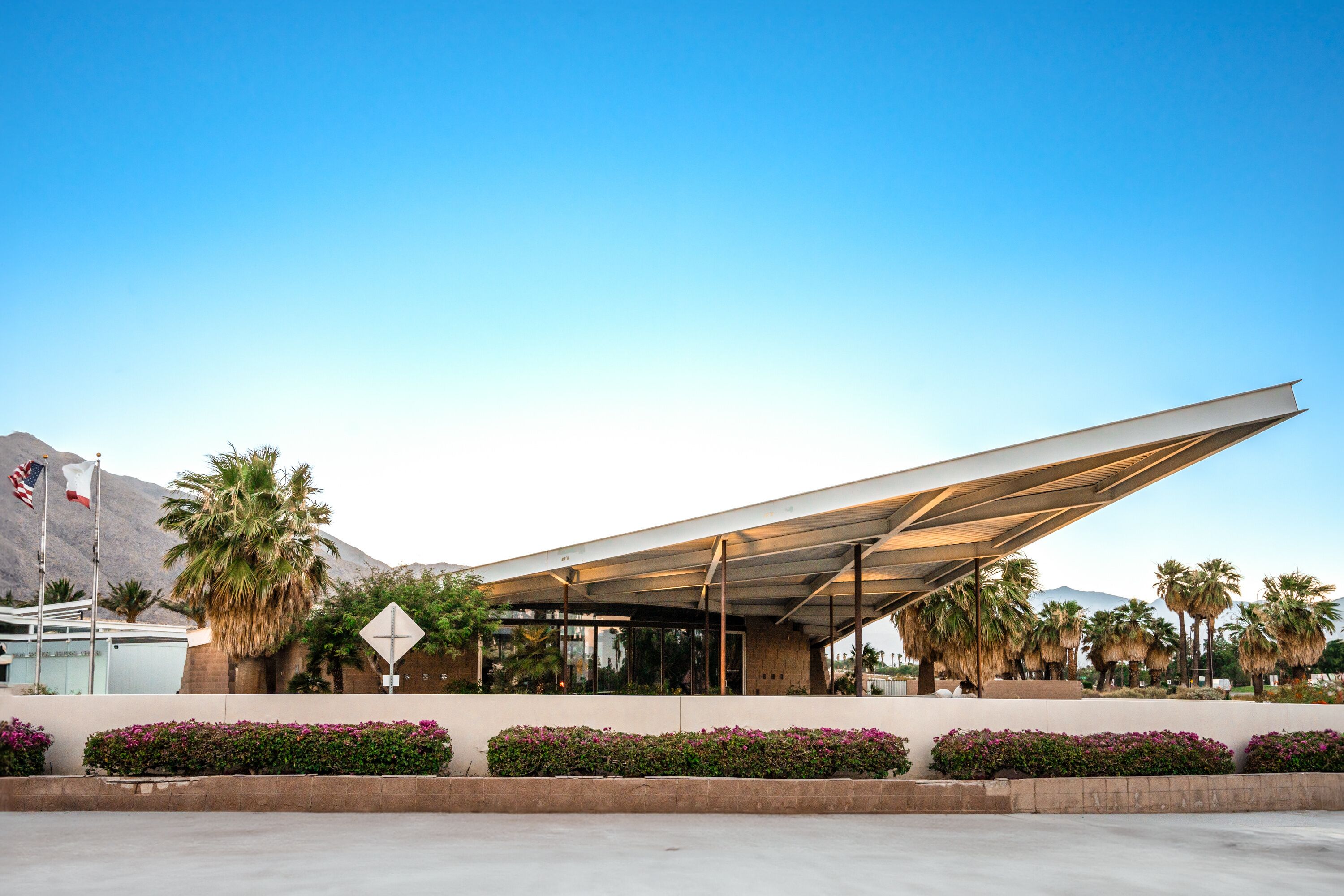 A modern building with a unique roof structure, palm trees, and a mountain in the background.