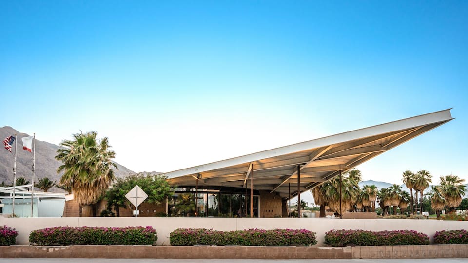 A modern building with a unique roof structure, palm trees, and a mountain in the background.